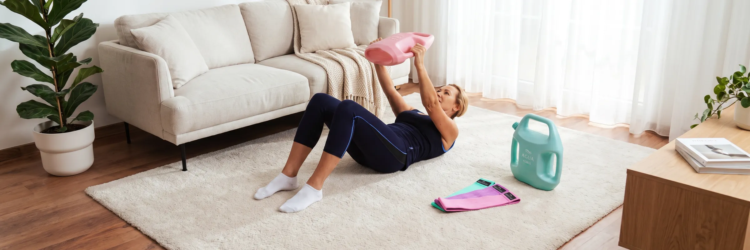 Woman lying on a cream rug in a bright living room, lifting a pink AguaForce water-filled weight overhead, with a mint AguaForce weight and resistance bands beside her and a sofa behind her